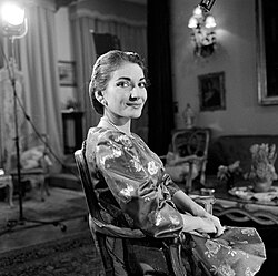 schwarz-weiß Foto von Maria Callas aus der TV-Show Small World, aus dem Jahr 1958. Maria Callas sitzend mit Hochsteckfrisur auf einem antiquaren verzierten Holzstuhl in einem langarmigen Kleid mit Blumenmuster, einer Perlenkette und Perlenohringen. Sie schaut nach rechts zum Fotografen und hat Ihre Arme auf den Lehnen des Stuhls auf ihrem Schoss zusammenhaltend liegen.