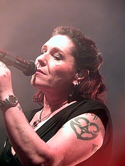 AnNa R. bei einem Konzert in Dresden mit der Band Silly 2022. AnNa R. hält ein Mikrofon. Sie hat auffällige Tätowierungen am linken Arm, darunter ein prominentes Design mit einem Herzen, das mit einem Pentagramm kombiniert ist. Sie trägt ein schwarzes ärmelloses Oberteil, eine Uhr am linken Handgelenk und eine Halskette mit einer Kette und einem Anhänger. Der Hintergrund ist rötlich neblig, vermutlich aufgrund von Bühnenbeleuchtung und Rauch der Bühneneffekte.