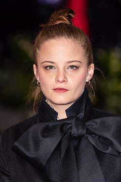 Jasna Fritzi Bauer auf der Berlinale 2020, trägt einen schwarzen Mantel mit großem Schleifenkragen, hat einen ernsten Gesichtsausdruck. Ein besonderes Detail ist die große, auffällige Schleife am Kragen, die aus einem glänzenden, satinartigen Material besteht und sehr sorgfältig gebunden ist. Die Frisur ist ein hoher Dutt, und als Accessoires trägt die Person kleine Kreolen-Ohrringe. Der Hintergrund ist unscharf, mit erkennbaren grünen Elementen – möglicherweise Pflanzen oder Bäume.