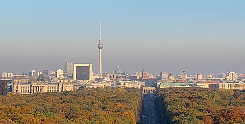 Farbfotografie von Berlin in der Vogelperspektive. Im Vordergrund stehen bewaldete Flächen vom Park Tiergarten und die Straße des 17. Juni führt zum Brandenburger Tor. Von links nach rechts sind das Reichstagsgebäude, der Fernsehturm, der Berliner Dom, das Rote Rathaus, das Brandenburger Tor und das Berliner Schloss zu sehen.