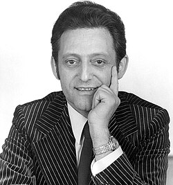 Autogrammkartenmotiv von Hans Rosenthal, der einen Nadelstreifenanzug und eine Krawatte trägt. Er lächelt und stützt eine Hand nachdenklich auf das Kinn, wobei die Armbanduhr am Handgelenk sichtbar ist. Das Bild ist in Schwarz-Weiß gehalten und vermittelt ein elegantes, formales Ambiente.