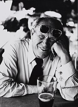 Gert Fröbe sitzt vor einem Bier und lacht in die Kamera, Sonnenbrille auf den Kopf geschoben