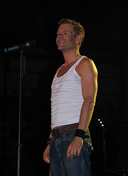 Peter Plate von Rosenstolz stehend auf der Bühne. Er trägt ein weißes Tanktop und eine blaue Jeans. Ein Gürtel ist um seine Taille gelegt, und er trägt ein schwarzes Armband am rechten sowie ein weiteres Armband am linken Handgelenk. Direkt neben Peter Plate steht ein Mikrofonständer mit einem befestigten Mikrofon. Der Hintergrund des Bildes ist dunkel gehalten und entstand während des Konzert in Leipzig 2006.