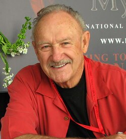Gene Hackman mit grauem Schnurrbart und kurzem grauen Haar nach hinten gekämmt. Trägt ein rotes Kurzarmhemd mit einem schwarzen T-shirt. Hintergrund erkennt man die Schrfitzüge NATIONAL und www. Rest wird von seinem Körper verdeckt.