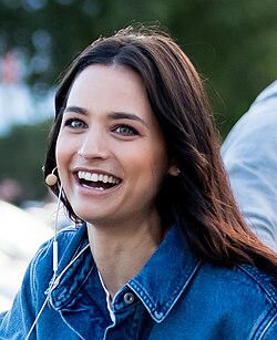 Amira Aly mit langen, braunen Haaren, die etwas über die Schultern fallen. Ihre Haare sind leicht wellig und wirken natürlich. Sie hat eine helle Hautfarbe und ihre Augen sind grünlich-blau. Ihre Augenbrauen sind gut geformt und ihre Wimpern sind deutlich sichtbar. Sie hat eine gerade Nase und einen vollen Mund; ihr Lächeln ist breit und offen, was einen sehr freundlichen und fröhlichen Ausdruck erzeugt. Ihre Gesichtszüge sind weich und harmonisch. Sie wirkt schlank und hat eine aufrechte Körperhaltung. Sie trägt eine Jeansjacke in einem mittelblauen Farbton, die offen über einem hellen Oberteil getragen wird (nur ein Teil des Unterteils ist sichtbar). Die Jeansjacke ist unkompliziert und lässig geschnitten. Sie trägt kleine, dezente Ohrringe; weitere Accessoires sind nicht sichtbar. Das Gesamtbild wirkt natürlich und ungezwungen.Amira Aly trägt weiße Ohrhörer, die sich in den Ohren befinden. Der Hintergrund des Bildes scheint im Freien zu sein, mit grünlicher Vegetation und einigen unscharfen Elementen, was auf eine natürliche Umgebung hinweist.